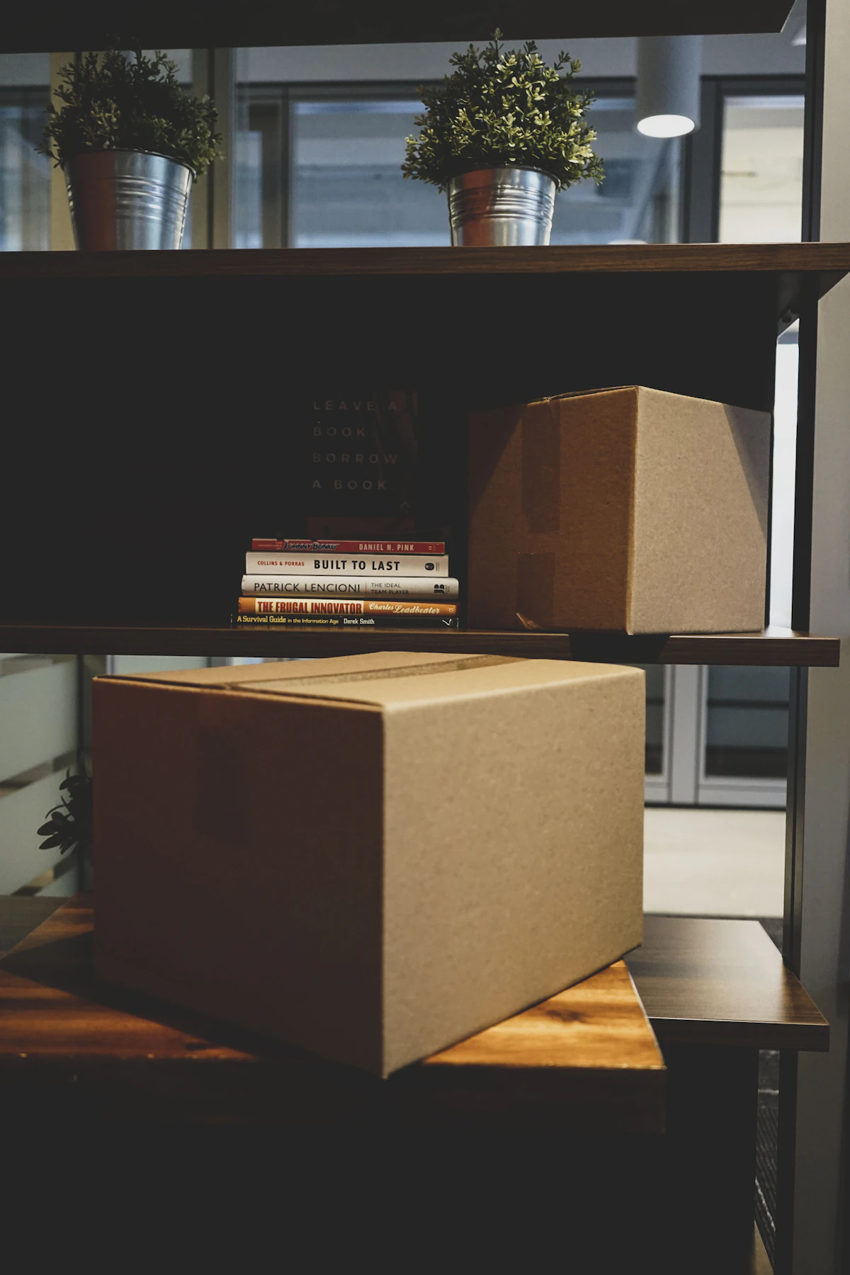 Packed cardboard boxes stacked on office shelving beside books and plants, ready for a move