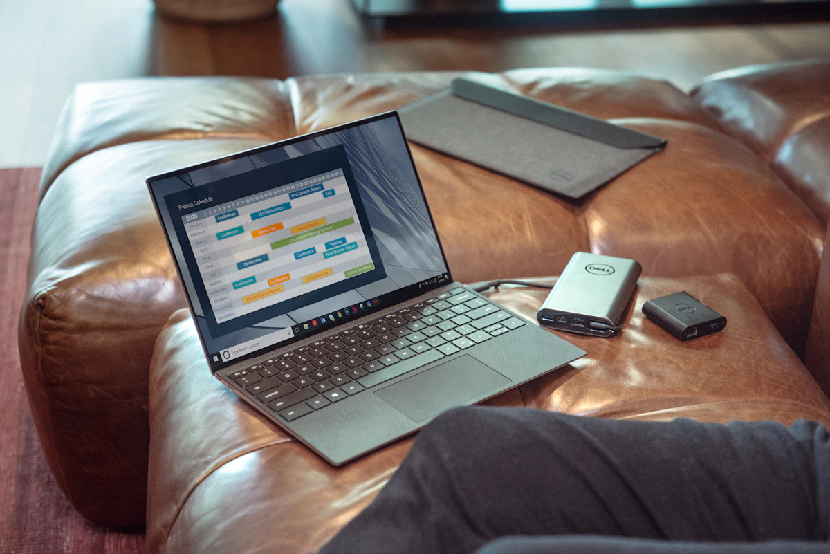 A laptop open on a leather ottoman showing a colour-coded schedule, beside a portable power bank